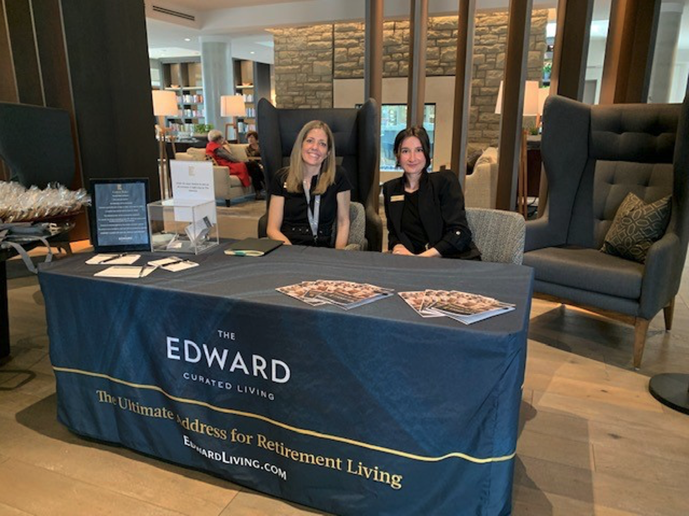 Two women sit at a promotional table with a tablecloth that says The Edward Curated Living The Ultimate address for retirement living edwardliving.com